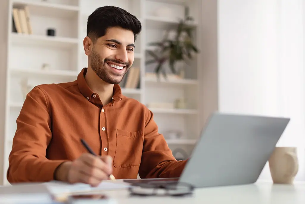 A man sits at a laptop smiling and making notes