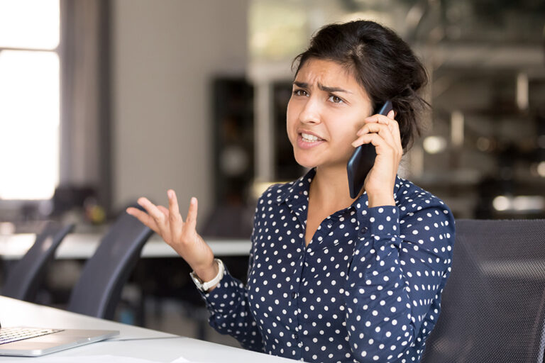 A woman sits at a desk while talking on the phone, making a concerned face and shrugging with her free hand in the air