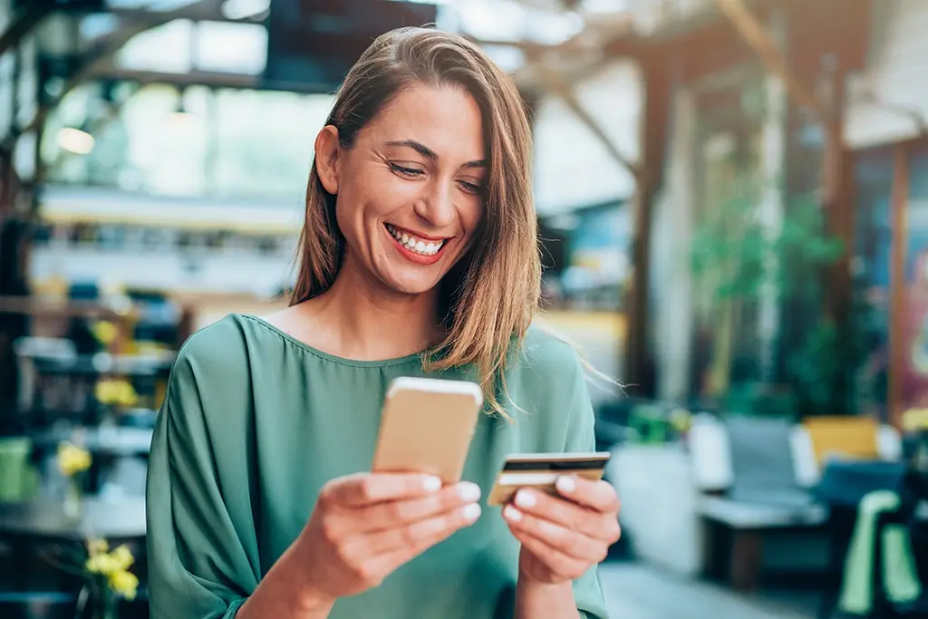 A woman smiles holding her credit card in one hand and her cell phone in another