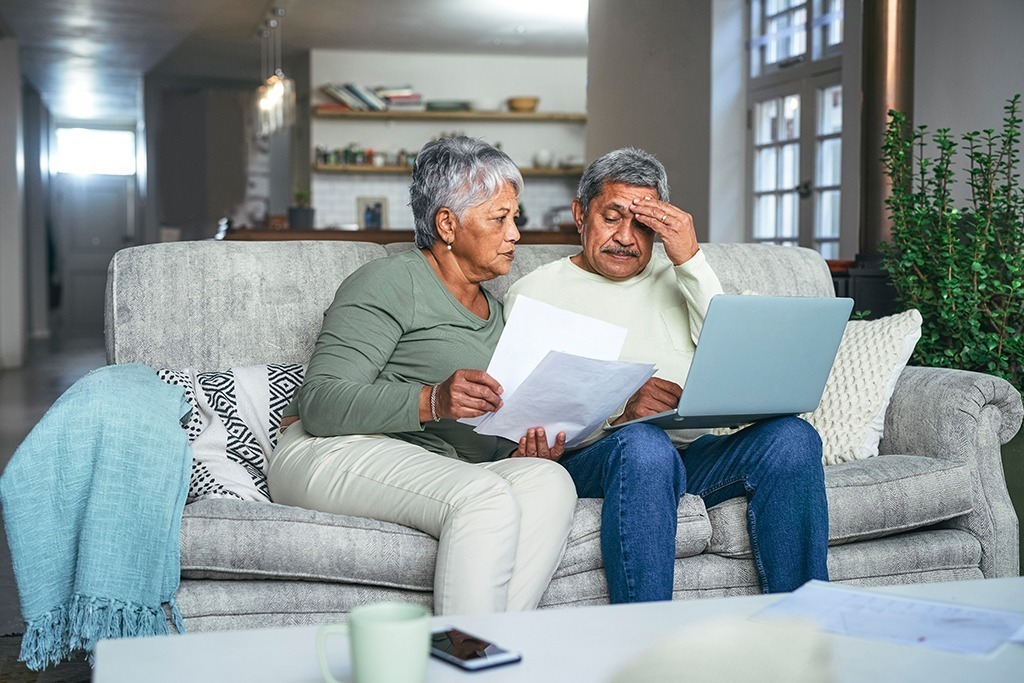 a couple in their 70s sits on a couch together, looking over paperwork while worried