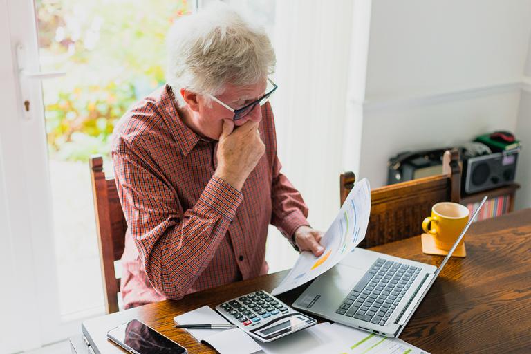 man in his 70s reviews financial papers while sitting at his laptop