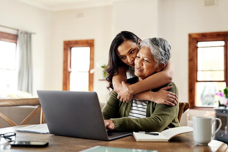 a woman in her 30s hugs a woman in her 70s as they look at a laptop together