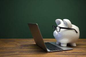 White Piggy Bank Side View Wearing Black Glasses Typing on Laptop with Black Chalkboard Background