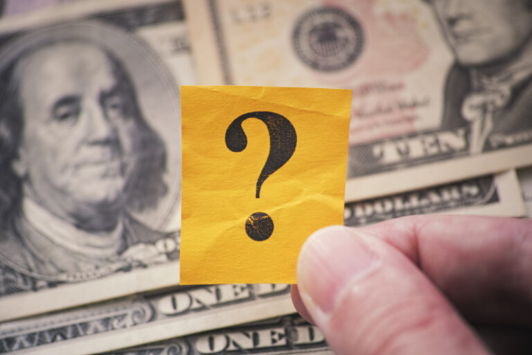 Person holds yellow paper note with question mark in his hand against US dollar background. Close up.