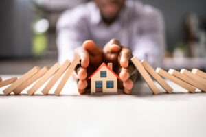 dominoes falling towards house from both sides, mans hand protecting house